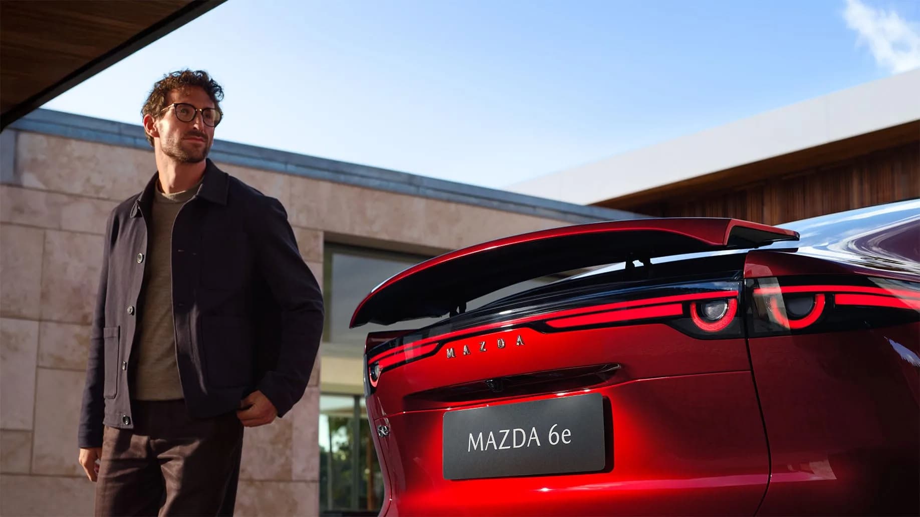 Mazda 6e — partea din spate a mașinii, cu eleronul ridicat, și un bărbat alături