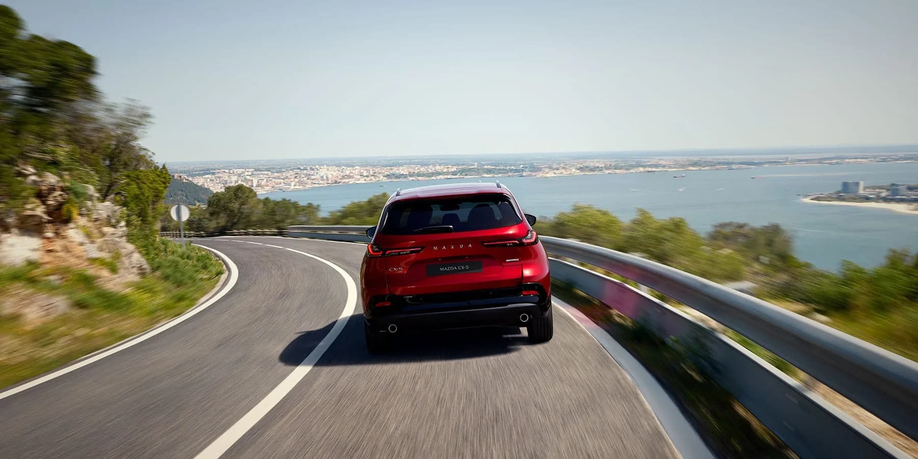 Mazda CX-5 roșie văzută din spate, rulând pe un drum de coastă