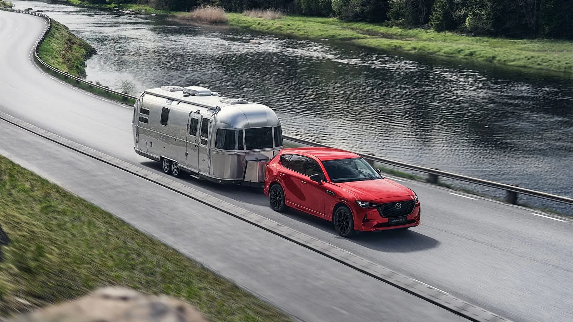 Mazda CX-60 roșie tractând o rulotă Airstream pe un drum de lângă un râu