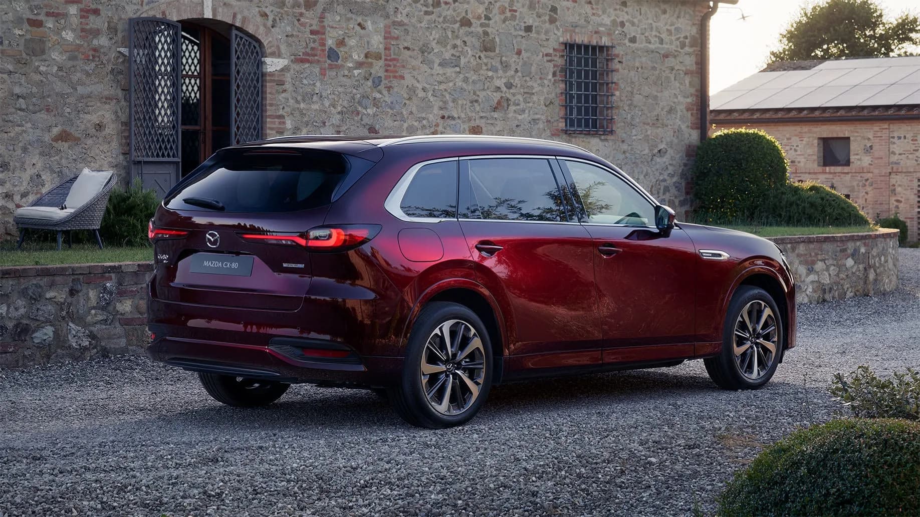Mazda CX-80 — CX-80 roșu-închis, din spate-lateral, lângă o casă de piatră