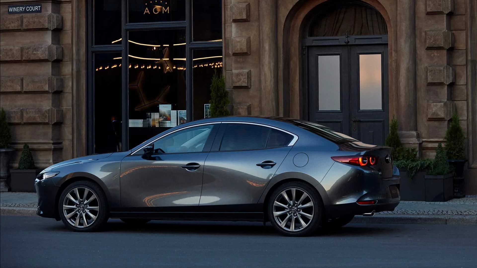 Mazda3 Sedan gri, văzută din lateral, parcată în fața unei clădiri